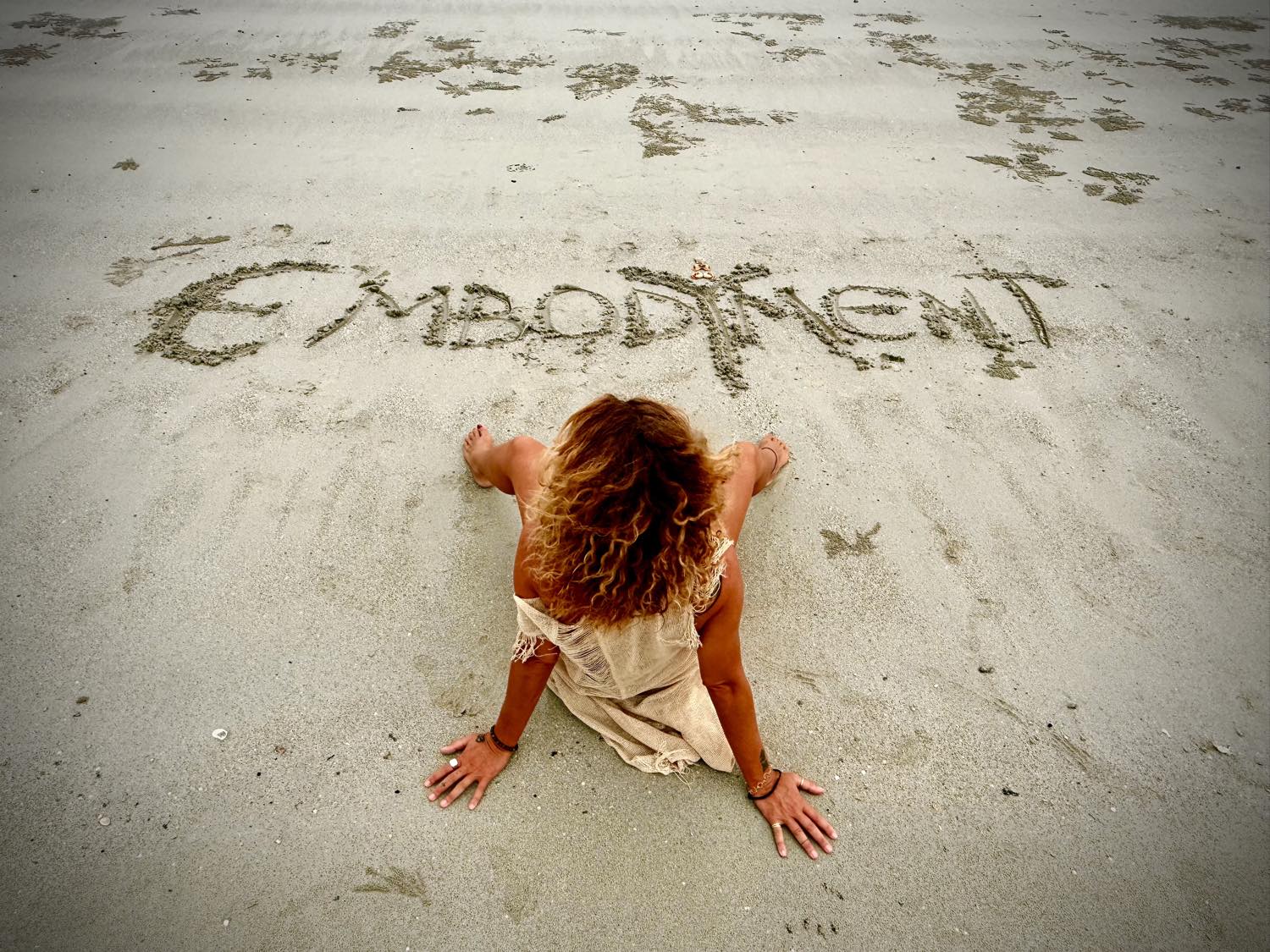 Eva Di Pierro sitting on the sand next to the word Embodiment written by hand